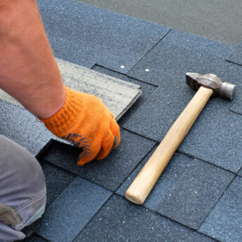 Contractor hands installing bitumen roof shingles using hammer in nails.